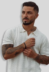 White short-sleeve polo shirt featuring textured zigzag patterns, with a classic collar. Model has tattoos visible on arms and wears a black bracelet.