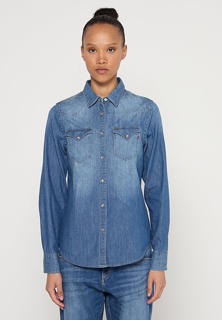 Camisa de mezclilla azul con mangas largas, cierre de botones en la parte frontal, dos bolsillos en el pecho y una textura ligeramente descolorida, con costuras en contraste.