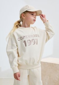 Sweat-shirt couleur crème avec "COPENHAGEN 1997" embossé dans une teinte plus foncée, associé à un pantalon doux assorti et une casquette beige.