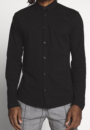 Hombre con camisa negra ajustada de manga larga con botones y pantalones grises de cuadros, mostrado desde el cuello hasta la mitad del muslo.