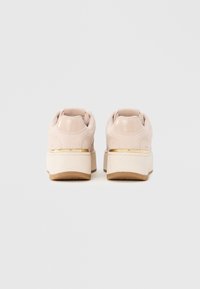 Zapatillas de plataforma beiges con suelas blancas gruesas, ribete metálico dorado sobre los talones y cuellos acolchados en los tobillos, vistas desde atrás.