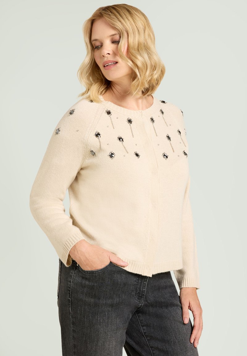 Hellbeiger Pullover mit dekorativen schwarzen Blumenverzierungen, Rundhalsausschnitt, langen Ärmeln und geradem Saum. Kombiniert mit dunklen Jeans.