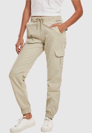 Beige cargobukser med elastiske manchetter og snørelukning i taljen, båret af en person i hvide sneakers og hvid cropped top, hånd i lommen.