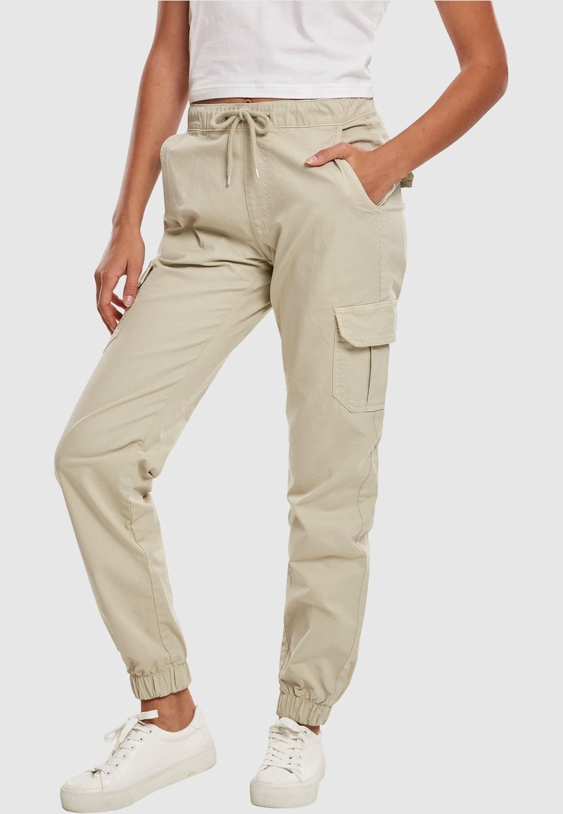 Pantalon cargo beige avec poignets élastiques et taille à cordon porté par une personne en baskets blanches et top court blanc, main dans la poche.