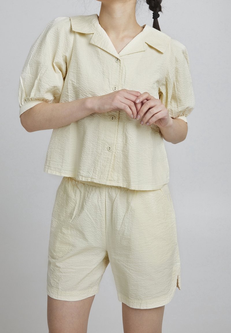 Femme portant un haut boutonné à manches courtes texturé jaune pâle avec un short assorti à taille élastique sur un fond uni.