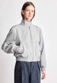 Grijze cropped bomberjack met hoge kraag, voorrits en geribbelde manchetten. Glad materiaal met subtiele textuur. Gepaard met getextureerde marineblauwe broek.
