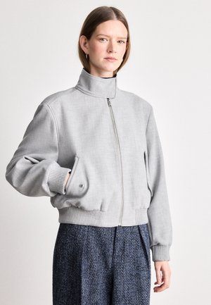 Grijze cropped bomberjack met hoge kraag, voorrits en geribbelde manchetten. Glad materiaal met subtiele textuur. Gepaard met getextureerde marineblauwe broek.