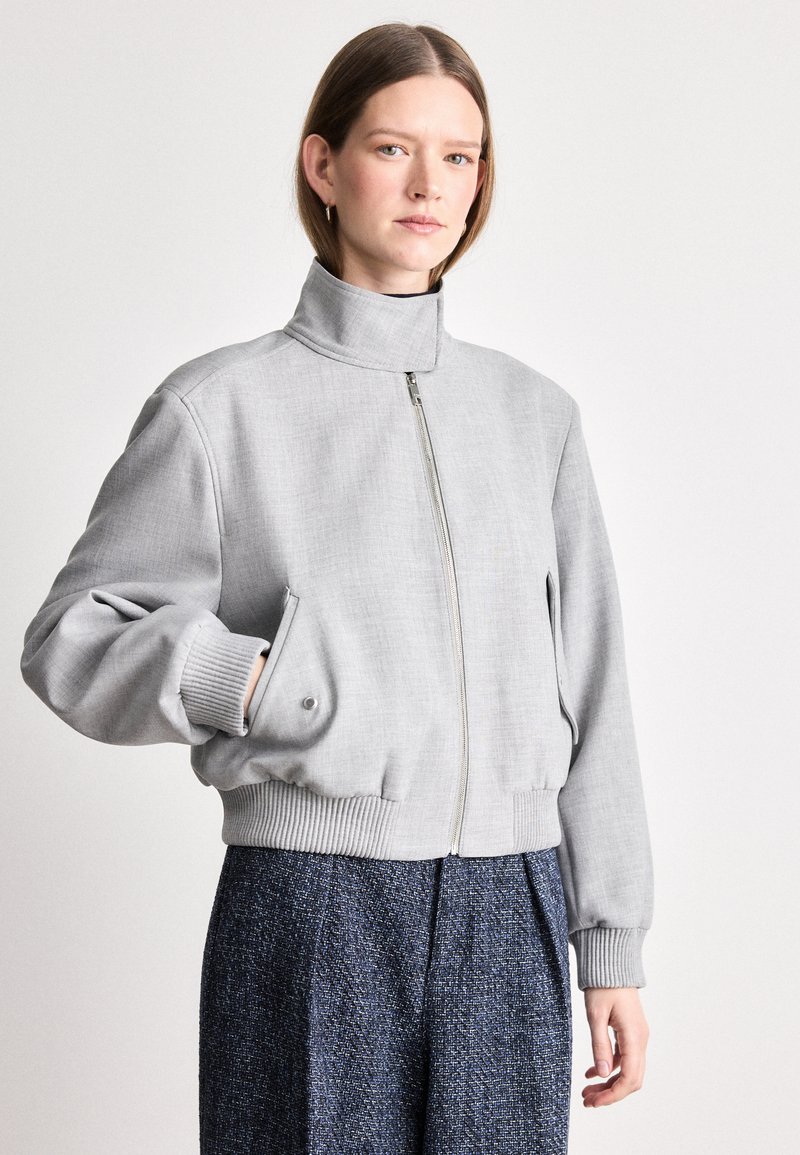Grijze cropped bomberjack met hoge kraag, voorrits en geribbelde manchetten. Glad materiaal met subtiele textuur. Gepaard met getextureerde marineblauwe broek.