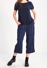 Navy blue top with short sleeves and pleated hem paired with cropped, navy striped pants. Model wears black shoes and socks.