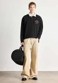 Μαύρο μακρυμάνικο πόλο πουκάμισο με λευκό γιακά και λογότυπο, συνδυασμένο με ελαφριά μπεζ φαρδιά παντελόνια, κρατώντας μια στρογγυλή μαύρη τσάντα.