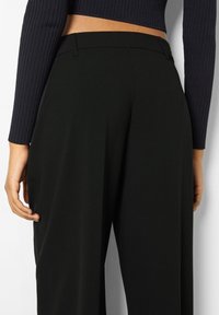 Black high-waisted trousers with a smooth, textured fabric; features side pockets and a tailored fit. The waistband is clean and discreet.