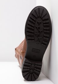 Botte en cuir marron avec semelle en caoutchouc noir montrant un motif de crantage et une fermeture éclair sur le côté, positionnée debout contre un mur et un sol blancs.