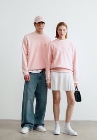 Δύο άτομα που φορούν ίδιες ανοιχτό ροζ μπλούζες· το ένα με φαρδιά τζιν παντελόνια και καπέλο, το άλλο με λευκή πλισέ φούστα κρατώντας μαύρη τσάντα χειρός.