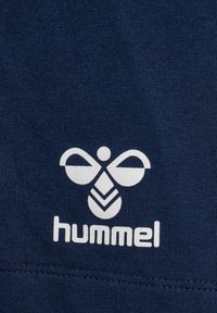 Tela azul marino con un logo blanco de Hummel que presenta un diseño simétrico que recuerda a un insecto y patrones de chevrón estilizados.