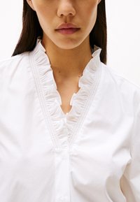 Blusa de algodón blanca con cuello volado, cierre de botones y tela texturada de forma sutil. Diseño simple con un aspecto limpio y clásico.
