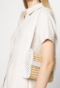 Una donna in una camicia a righe chiara tiene in mano una borsa intrecciata decorata con perline bianche, che mostra un design elegante e testurizzato.