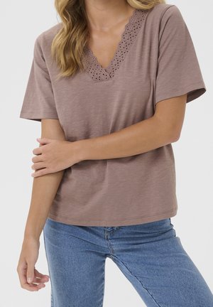 Frau trägt ein hellbraunes T-Shirt mit V-Ausschnitt und Spitzenbesatz sowie blaue Jeans, posiert mit einem Arm vor dem Körper verschränkt vor einfachem Hintergrund.