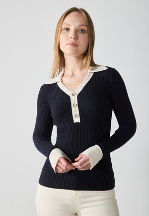 Navy ribbed knit sweater with a cream collar, button placket featuring three gold buttons, and cream cuffs with thumbholes.