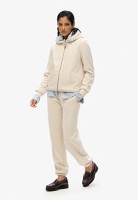 Felpa beige con cappuccio, zip, tasche a canguro e cordoncino. Abbinata a pantaloni jogger coordinati e una maglietta a righe sotto.