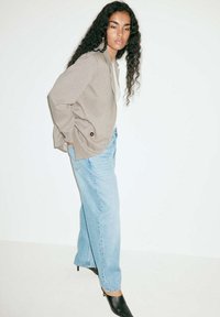 Femme aux longs cheveux bouclés portant un blazer à carreaux, un jean bleu clair et des chaussures à talons noires, debout devant un fond blanc.