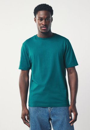 Giovane uomo nero in piedi, indossa una maglietta verde acqua a maniche corte e jeans blu su uno sfondo chiaro e uniforme.