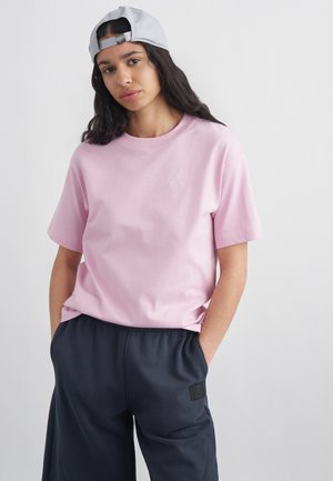 Młoda kobieta w jasnoróżowej koszulce, czarnych spodniach i białej czapce z daszkiem noszonej do tyłu, z rękami w kieszeniach.