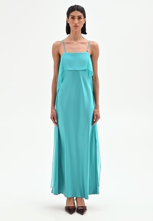 Femme aux cheveux foncés portant une robe maxi turquoise sans manches avec des bretelles fines et des talons pointus foncés, debout sur un fond blanc.