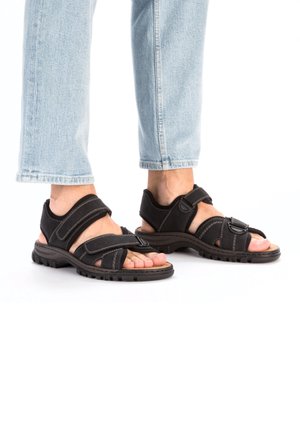 Schwarze Sandalen mit gepolsterten Riemen, ausgestattet mit zwei verstellbaren Klettverschlüssen. Dicke Gummisohle mit strukturiertem Profil für besseren Halt.