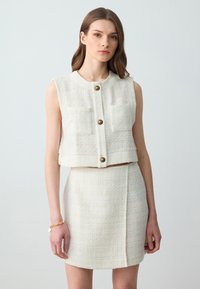 Witte tweed crop vest met vier voorzakken, gouden knoopsluitingen, gecombineerd met een bijpassende mini skirt met een naaddetail aan de zijkant.