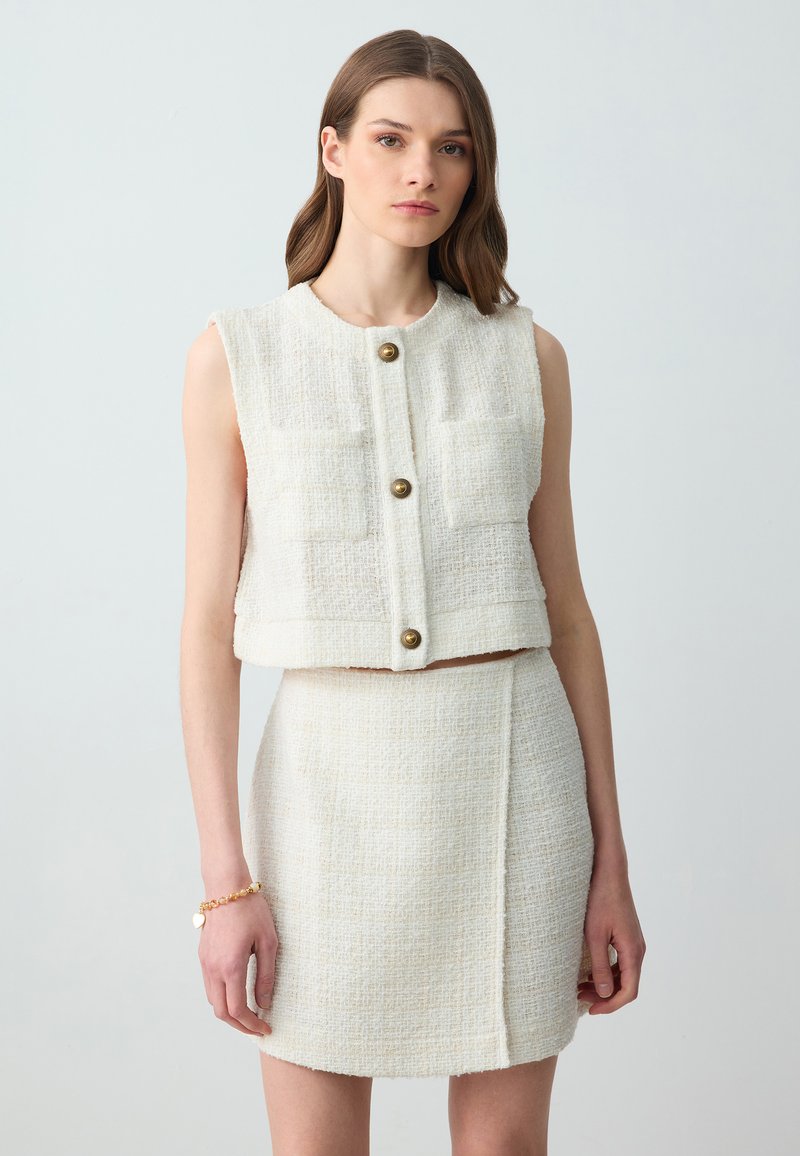 Witte tweed crop vest met vier voorzakken, gouden knoopsluitingen, gecombineerd met een bijpassende mini skirt met een naaddetail aan de zijkant.