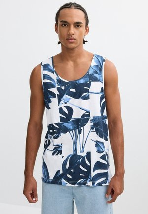 Joven con cabello trenzado que lleva una camiseta blanca sin mangas con grandes patrones de hojas tropicales en azul oscuro y vaqueros azul claro.
