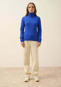Strickrollkragenpullover in lebhaftem Blau, gerippte Textur, kombiniert mit hellbeigen Hosen und cremefarbenen Sneakers vor einem neutralen Hintergrund.
