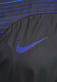 La tela negra presenta franjas azules horizontales y el logo de Nike en color púrpura. La textura parece suave y ligera.