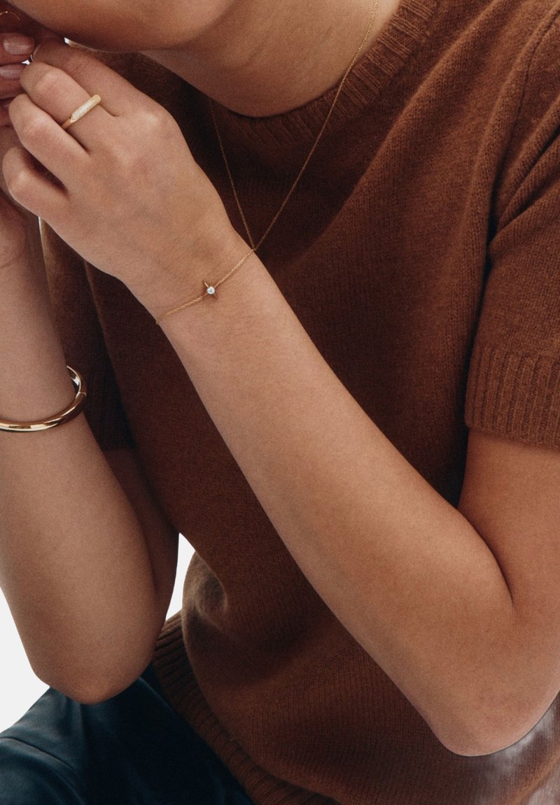 Gold bracelet with a small gem, textured gold ring, and a fitted brown knitted sweater with short sleeves. Soft, smooth skin tones.