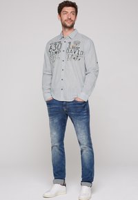 Camisa abotonada de color gris claro con gráficos de texto, combinada con jeans azules y zapatillas blancas. La camisa tiene un collar y mangas largas.