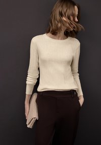 Maglione beige chiaro a coste con maniche lunghe abbinato a pantaloni marrone scuro. La modella tiene una pochette beige intrecciata e texturizzata. Sfondo neutro.