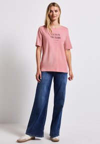 Růžové tričko s textem "CALM ON THE INSIDE" v černé barvě, v kombinaci s volnými modrými džíny a krémovými teniskami. Neformální design.