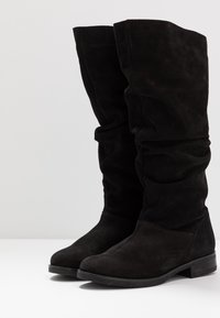 Czarne zamszowe botki do połowy łydki z niskimi obcasami blokowymi i lekko opadającymi cholewkami, pokazane obok siebie na białej powierzchni.