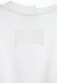 Camiseta blanca con cuello redondo, pequeña abertura en la parte posterior y etiqueta de cuero blanco con texto en relieve. Tela lisa y sin textura.