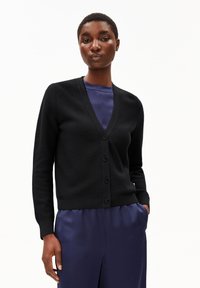 Cardigan nero a maglia con scollo a V, dotato di chiusura con bottoni, maniche lunghe e polsini a costine. Indossato sopra una maglietta blu.