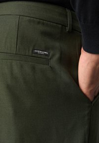 Pantalons vert olive en tissu texturé, dotés d'une poche arrière avec une étiquette noire portant la mention "SCOTCH & SODA, AMSTERDAM."