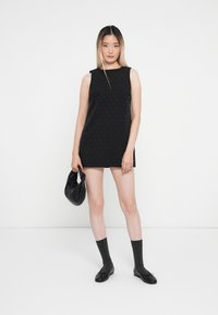 Rochie mini fără mâneci, de culoare neagră, cu un model cu puncte, asortată cu șosete gri până la genunchi și pantofi negri. Modelul ține o geantă mică neagră.