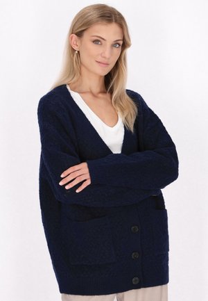 Donkerblauwe gebreide cardigan met een V-hals, grote voorkruiszakken en knoopsluiting. Beschikt over een gewatteerd patroon en een losse pasvorm.