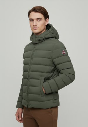 Muž v olivově zelené prošívané bundě s kapucí a bočními kapsami stojí před jednobarevným světle šedým pozadím.