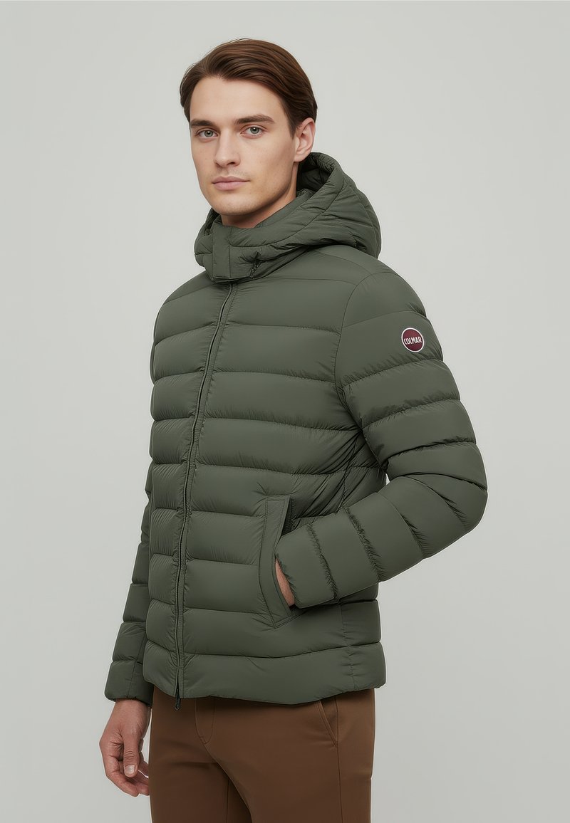 Hombre con chaqueta acolchada verde oliva con capucha y bolsillos laterales, de pie contra un fondo gris claro liso.