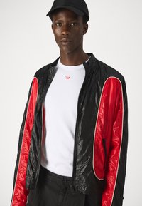 Sort og rød vindjakke med lynlås foran og kontrasterende hvid kant. Bæres over en hvid t-shirt med en simpel rund hals.