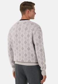 Man som bär en ljusgrå mönstrad tröja och mörkgrå byxor, sedd bakifrån, tittar åt höger mot en vit bakgrund.