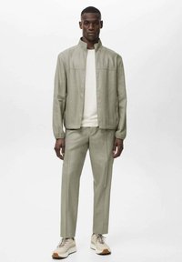Lichtgroene jas met hoge kraag, gecombineerd met bijpassende broeken. De outfit heeft een soepele textuur en een minimalistisch ontwerp, aangevuld met beige sneakers.