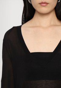 Musta V-kaelusega top, läbipaistva kangaga, pikkade varrukatega ja kihilise disainiga musta saki kohal, esitleb kergelt tekstuuriga kangast.
