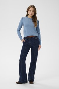 Nő áll zsebekkel a kezében, kék hosszú ujjú pulóvert gyöngy részletekkel és sötét szárú, bővülő farmert visel barna cipővel.